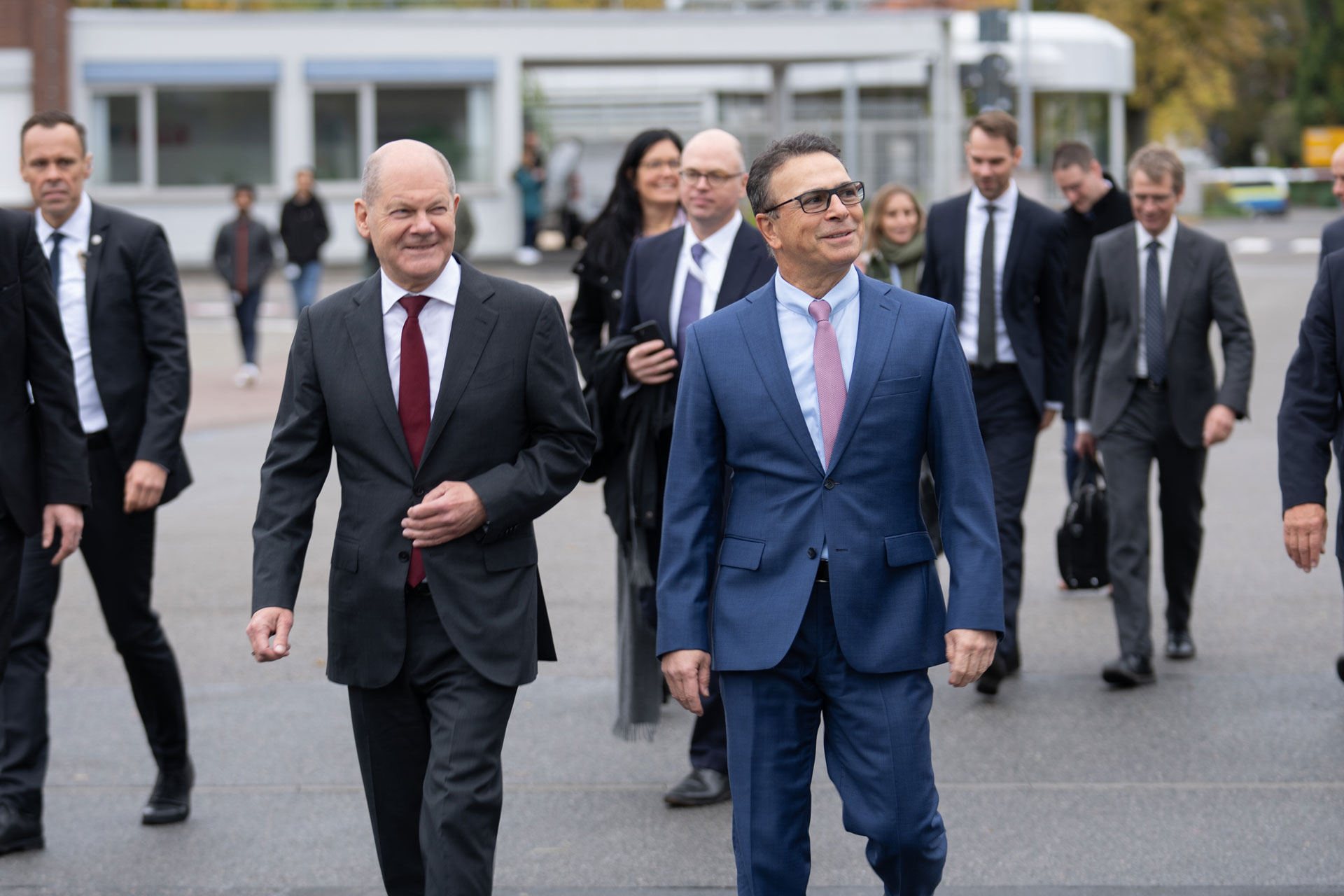 The picture shows two men walking in the foreground, looking to the left and smiling. Other people can be seen in the background.