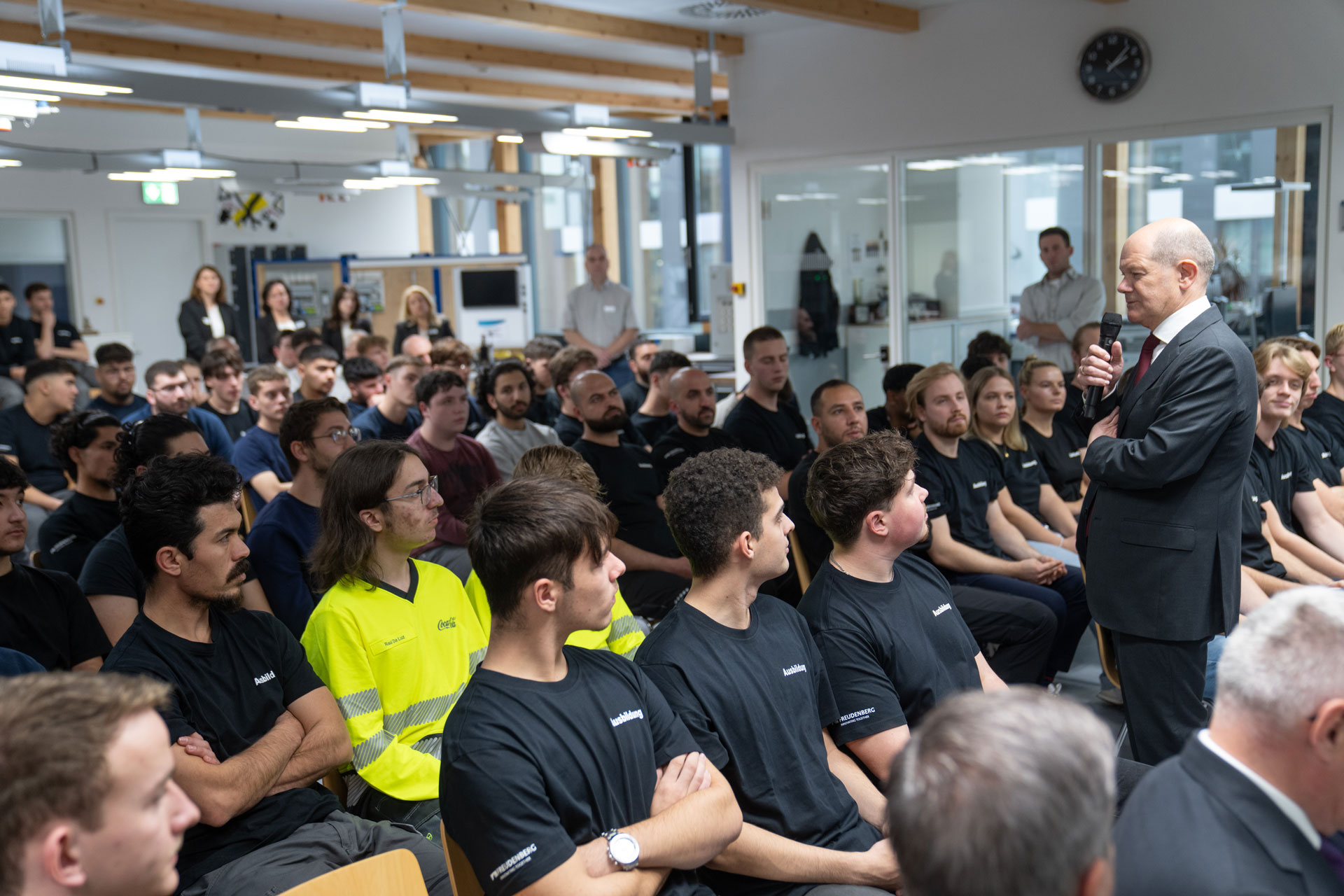 The picture shows a man holding a microphone and standing in front of a group of people sitting in rows looking at the man