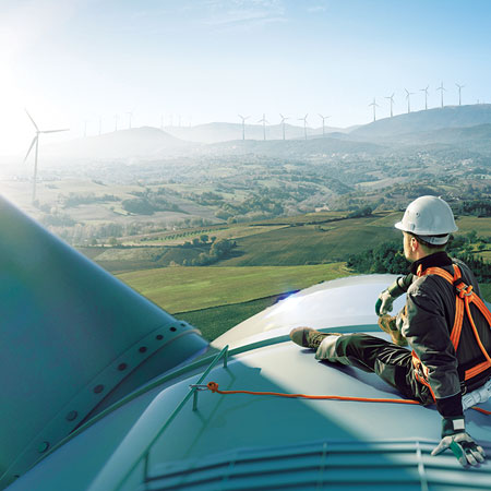 Erfahren Sie mehr über unsere nachhaltigen Aktivitäten Ein Mann, der Sicherheitsgurte und einen Helm trägt, sitzt auf einer Windkraftanlage und blickt in Richtung eines verschwommenen Horizonts
