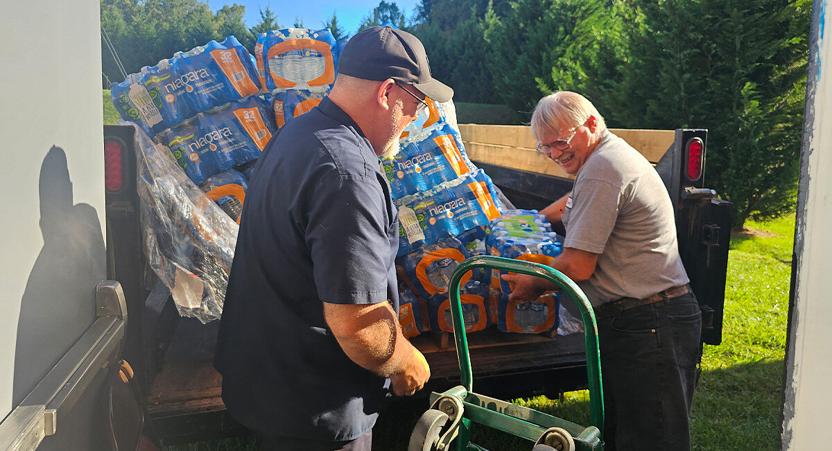 Das Bild zeigt zwei ältere Männer beim Entladen von Wasserflaschen aus einem Lastwagen