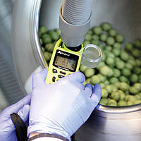 behandschuhte Hände halten ein Messgerät in einem runden Stahlbehälter mit grünen Dragees