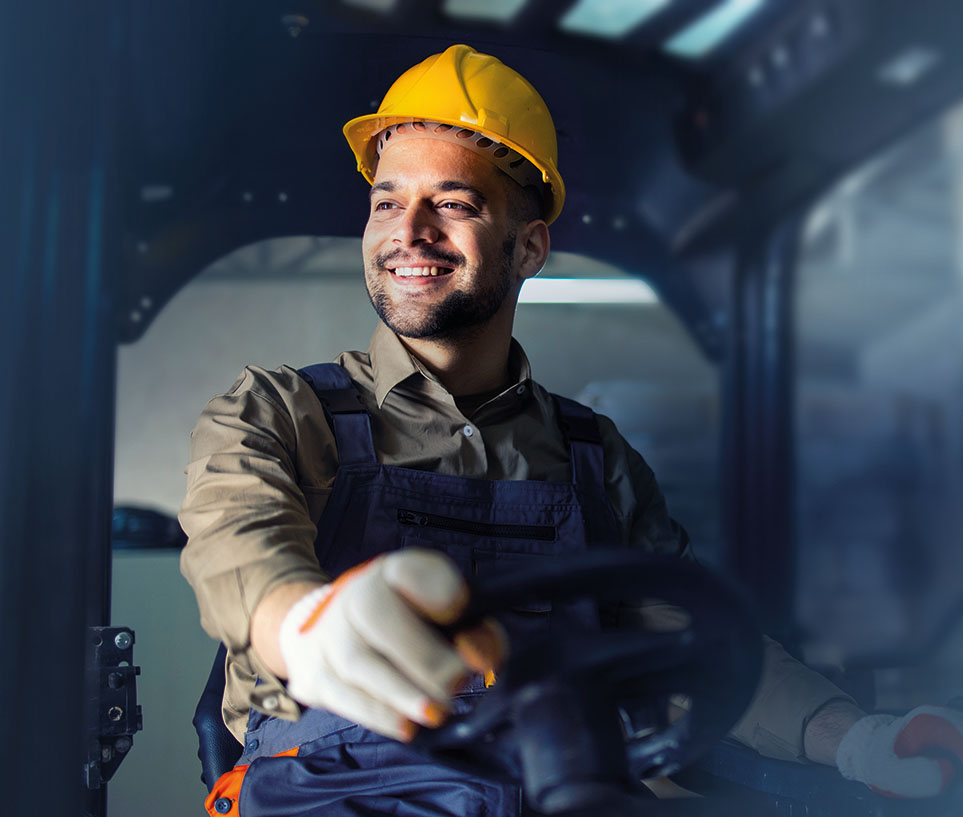 Young male in working uniform and yellow hardhat operating forklift machine in warehouse storage room.